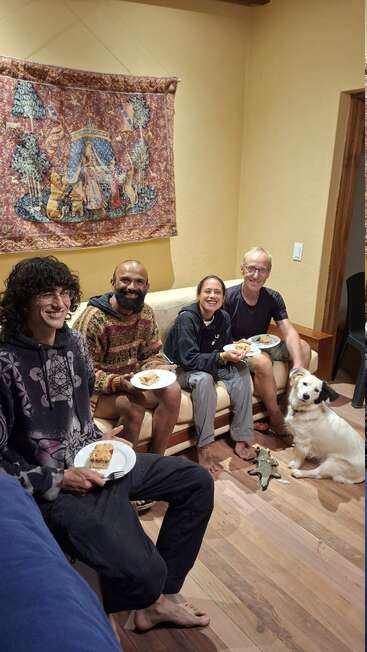 Four people sit together in a cozy room, smiling and enjoying food. A white dog sits on the floor, surrounded by toys. Tapestry decorates the wall.