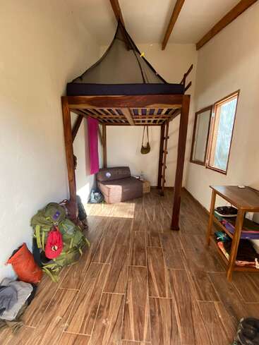 A simple, tidy room with wooden floor, lofted bed with mosquito net, open window, backpack on the floor, shelving unit, and bags and boxes in corner.