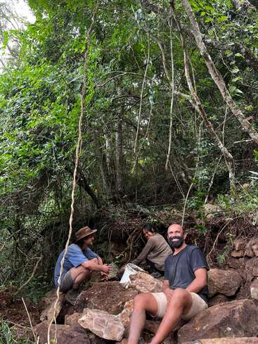 Three people sit among rocks and dense forest vegetation, smiling and relaxing. They appear to be working or exploring together in a natural, outdoor environment.