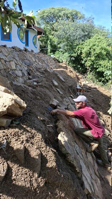 A man in a red shirt and cap builds a stone retaining wall on a sloped hillside near a white house surrounded by lush green trees.