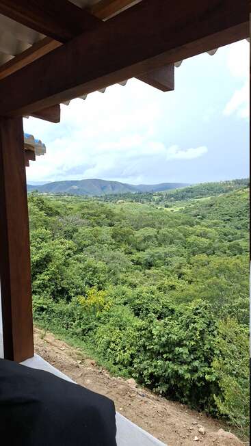 A wooden porch overlooks a lush, green forest with rolling hills in the distance. The sky is cloudy, creating a peaceful, serene, and natural atmosphere.