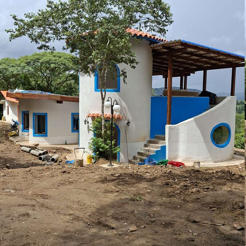 This image shows a unique, white and blue house with rounded walls, a rooftop terrace, and outdoor stairs. Surrounded by trees, some construction materials remain outside.