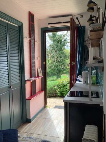 Sunlit tiny house interior features a kitchenette, green-trimmed door, chalkboard, and shelves. The open door reveals lush greenery and a peaceful outdoor scene.