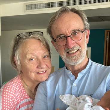 Una pareja de ancianos sonríe afectuosamente, de pie, muy juntos, en un interior. El hombre sostiene con delicadeza a un recién nacido envuelto en una manta, captando un tierno momento familiar.