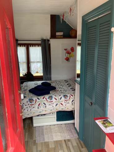 This cozy bedroom features a colorful floral bedspread, blue closet doors, wooden flooring, red door, bunting, books, artwork, and natural light through curtained windows.
