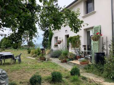 A charming countryside house with light walls, green shutters, and many potted plants. There's a cozy patio, garden furniture, and lush greenery under an overcast sky.