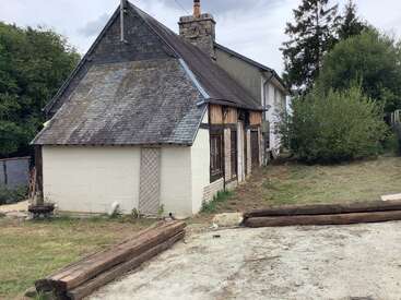 This image shows an old house with a slanted slate roof, white walls, wooden beams, surrounded by greenery and a partially completed yard with timber edging.