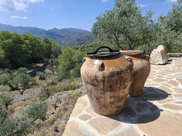 Dos grandes macetas de barro rústico se sientan en un patio de piedra bajo el sol, con vistas a un pintoresco valle con olivos y montañas lejanas bajo un cielo azul.