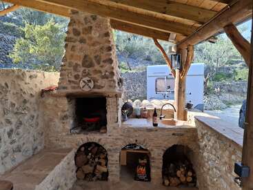 Esta imagen muestra una cocina rústica al aire libre con horno de piedra, almacén de leña, fregadero y techo de madera cubierto. Fuera hay una autocaravana.