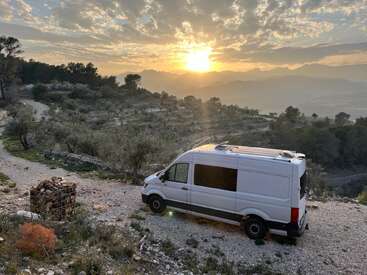 Ein weißer Lieferwagen parkt auf einem Schotterweg mit Blick auf terrassenförmige Hügel, Bäume und Berge. Die Sonne geht unter und wirft ein goldenes Licht durch einen teilweise bewölkten Himmel.