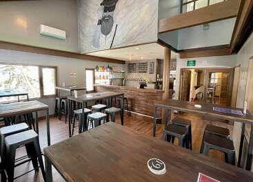 This cozy bar features wooden high tables and stools, a rustic counter, drink selections, bright windows, a mezzanine, and wall art of a bearded man.