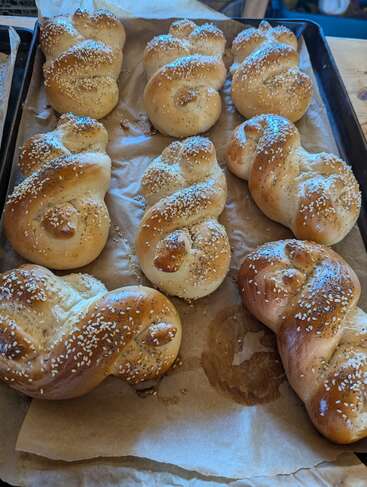 Auf einem Backblech liegen mehrere frisch gebackene, goldbraune Flechtbrötchen. Mit Sesam bestreut ruhen sie auf Pergamentpapier und sind bereit, genossen zu werden.