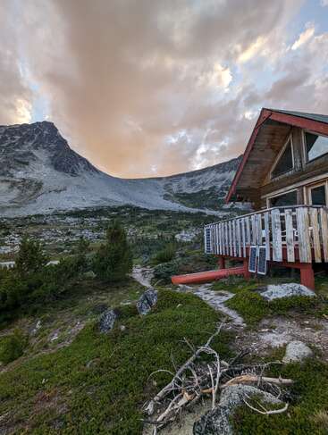 Eine rustikale Holzhütte steht bei Sonnenuntergang an einem Berghang, umgeben von Grün und felsigem Gelände, mit dramatischen Wolken unter einem friedlichen Himmel.