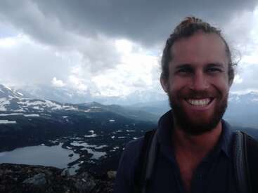 Ein Mann mit Bart und langen Haaren steht im Freien und lächelt breit. Im Hintergrund sind Berge, Seen und Schneeflecken unter einem bewölkten Himmel zu sehen.