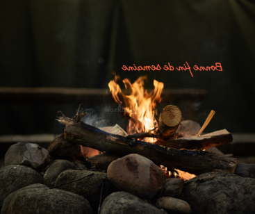 Das Bild zeigt ein Lagerfeuer mit Holzscheiten und Steinen und den Text \"Bonne fin de semaine\" in orangefarbener Kursivschrift oben auf einem dunklen Hintergrund.
