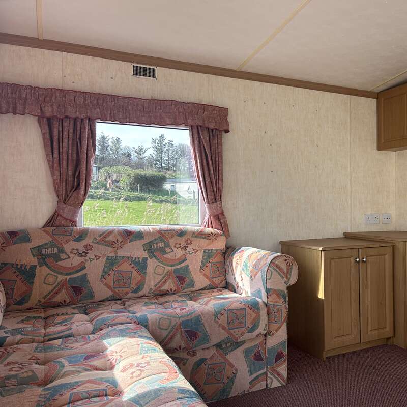 Esta imagen muestra el interior de una caravana con un sofá estampado, armarios de madera y una ventana con cortinas rosas, con vistas a una zona de césped en el exterior.