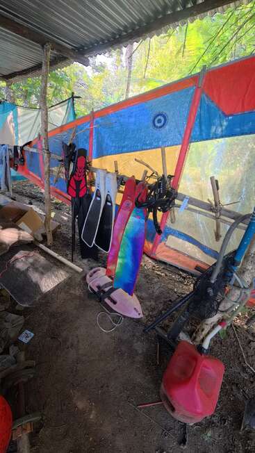 La imagen muestra un refugio rústico al aire libre con equipo de buceo o snorkel, aletas de colores, trajes de neopreno, tubos de buceo y un bidón de gasolina rojo sobre un suelo de tierra.