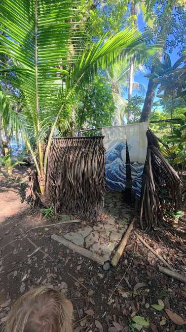 Una pequeña cabaña rústica construida con hojas de palmera se encuentra en medio de frondosos árboles tropicales, con una cortina que reproduce el grabado «La gran ola» de Hokusai a modo de entrada.