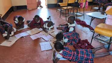 Eine Gruppe junger Mädchen in karierten Uniformen liegt auf dem Boden des Klassenzimmers und arbeitet gemeinsam an ihren Aufgaben, umgeben von Heften, Stiften und verstreuten Papieren.