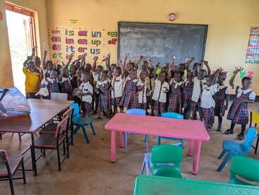 Eine fröhliche Gruppe junger Schulkinder in Uniformen hebt enthusiastisch die Hände in einem lebhaften Klassenzimmer, umgeben von bunten Tischen, Stühlen und lehrreichen Wanddekorationen.