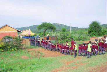 Das Bild zeigt eine Gruppe von Kindern in roten Uniformen, die vor einem Schulgebäude stehen, mit einem gelben Zelt und einer bergigen Landschaft im Hintergrund.