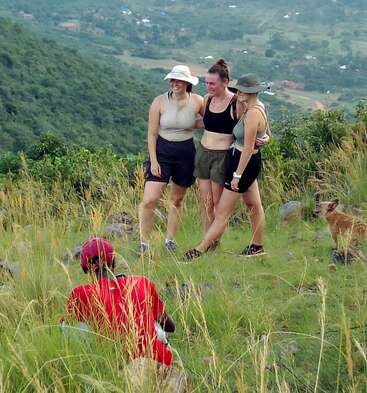 Das Bild zeigt drei Frauen in Sportkleidung, die über eine Wiese laufen. Im Vordergrund kniet ein Mann in einem roten Hemd und in der Nähe steht ein Hund.