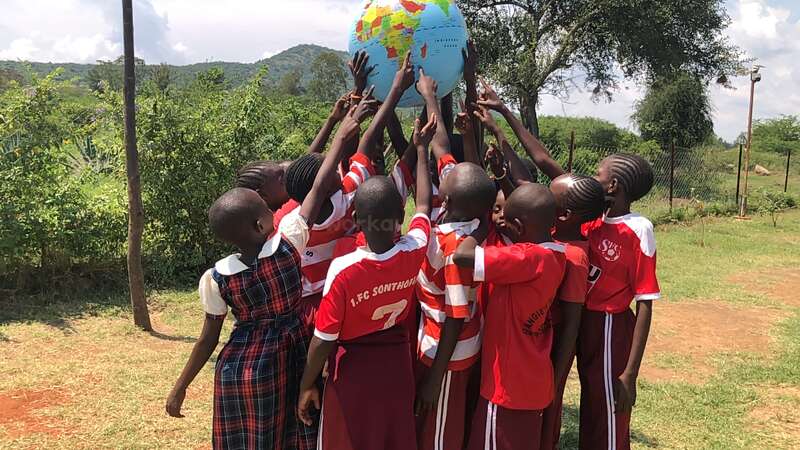 Eine Gruppe von Kindern in roten Uniformen versammelt sich im Freien und hebt gemeinsam eine große aufblasbare Weltkugel über ihre Köpfe, die Einheit und globale Verbindung symbolisiert, umgeben von Grün.
