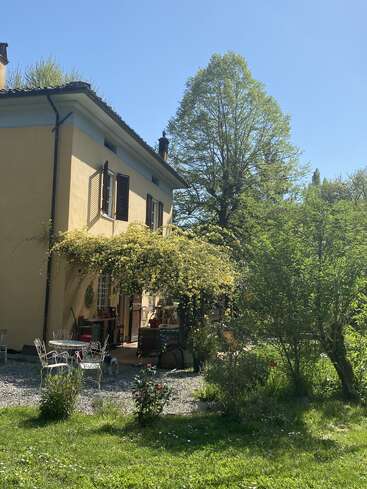 Ein charmantes gelbes Haus mit offenen Fensterläden ist umgeben von üppigem Grün, blühenden Blumen, Sitzgelegenheiten im Freien und einem kleinen Hund, der sich im hellen Sonnenlicht sonnt.