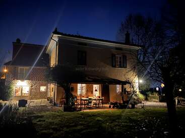 Ein gemütliches Haus leuchtet warm in der Nacht, umgeben von Bäumen und einem grasbewachsenen Hof. Sitzgelegenheiten im Freien schaffen eine einladende Atmosphäre unter dem dunklen, klaren Abendhimmel.