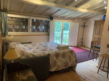 Dieses gemütliche Schlafzimmer verfügt über eine geblümte Tagesdecke, Holzböden, große Fenster mit Blick auf den Garten, rustikale Möbel, einen Kleiderständer und eine ruhige, einladende Atmosphäre mit natürlichem Licht.