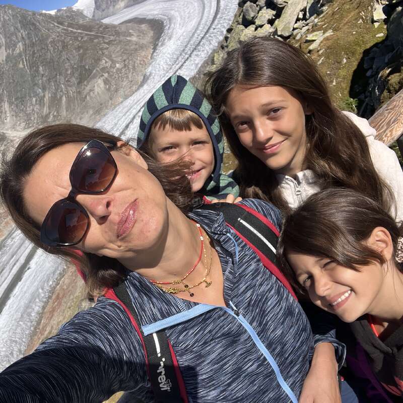 Eine Frau und drei Kinder lächeln für ein Selfie im Freien, mit einem dramatischen Gletscher und schroffen Bergen im sonnigen Hintergrund, die alle glücklich und abenteuerlustig aussehen.