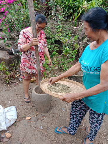 Two women are processing coffee beans outdoors. One woman is pounding the beans with a wooden pestle, while the other holds a woven tray of beans.
