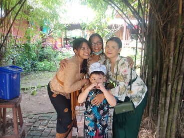 Three women in traditional attire and one young boy pose together outdoors, smiling happily. Lush green plants and a village-like wooden house form the background scene.