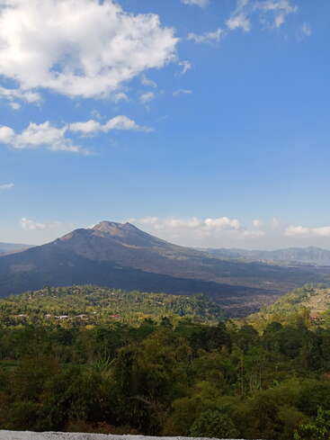 This image features a majestic mountain under a bright blue sky with scattered clouds, lush green forest, and a small village nestled at its base.