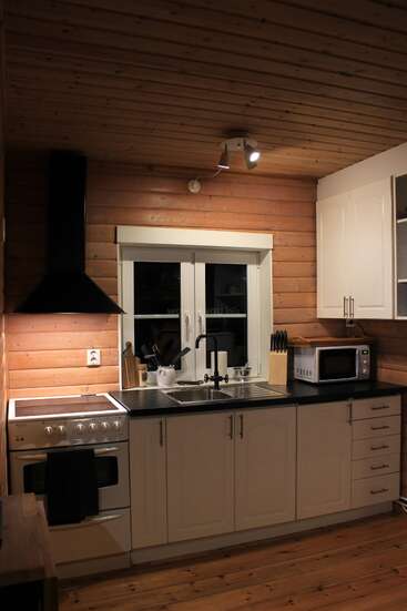 Diese gemütliche Küche hat Holzwände und -decken, weiße Schränke, einen Elektroherd, schwarze Arbeitsflächen, eine Mikrowelle, eine Spüle, Geschirr, Schneidebretter und eine Deckenbeleuchtung.