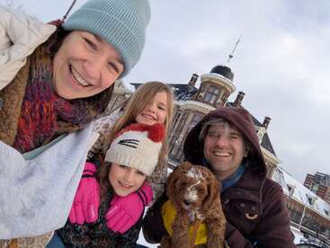 A happy family of four, bundled in winter clothing, poses outdoors on a snowy day with their adorable brown puppy. A beautiful old building is behind them.