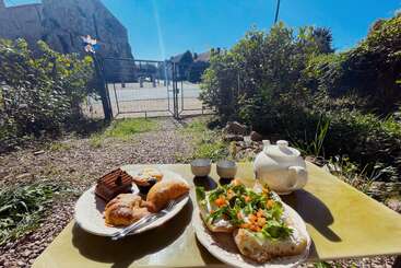 O café da manhã é servido ao ar livre em um dia ensolarado. Os pratos têm doces, bolos, pão com vegetais, acompanhados por um bule e xícaras. A cena é cercada por vegetação e prédios.