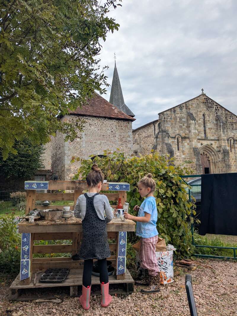 Two children play at a rustic outdoor mud kitchen, with a historic stone church and lush greenery in the background, creating a peaceful, countryside scene.
