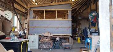 A workshop with a partially constructed wooden structure in the center, surrounded by stacks of wood, workbenches, tools, cardboard, and various materials and equipment.