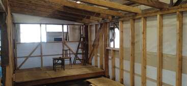 This image shows a wooden structure under construction, featuring exposed framing and beams, a small platform, a wooden chair, and a ladder near large windows.
