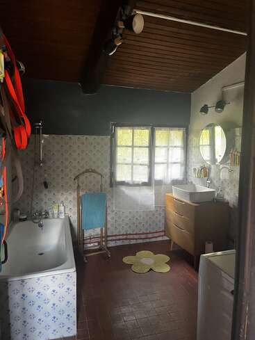 Este cuarto de baño cuenta con bañera, azulejos vintage, techo de madera, lavabo tipo cómoda, espejo circular, alfombra en forma de flor, toallero, lavadora y luz natural desde la ventana.