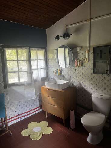 Este cuarto de baño presenta una pared con motivos florales, tocador de madera con lavabo, espejo circular, alfombra en forma de flor, inodoro, ventana con cortinas transparentes y techo de madera oscura. Ambiente acogedor.