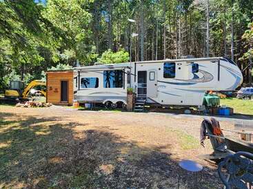 A large white RV with slide-outs sits in a wooded clearing beside a small wooden shed, surrounded by trees, outdoor supplies, and construction equipment.