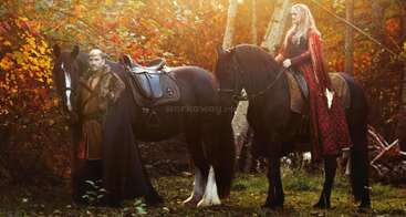 A man and a woman in medieval-style clothing stand with two black horses in a vibrant autumn forest. Sunlight filters through the colorful leaves dramatically.