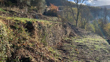 A sunlit terraced hillside features a rustic stone retaining wall, sparse vegetation, leafless trees, and distant houses, blending naturally with the surrounding autumn landscape and mountains.