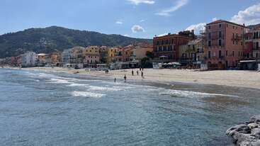 A picturesque seaside town with colorful buildings lines a sandy beach. Gentle waves lap ashore, people stroll, and hills rise in the background under blue skies.
