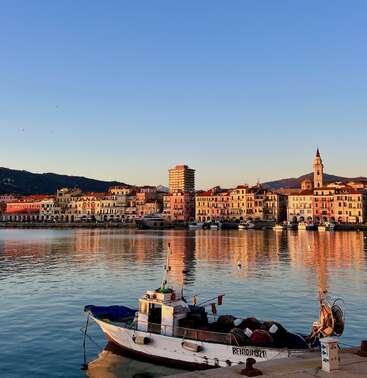A small fishing boat floats in a peaceful harbor, with colorful buildings and a church tower along the waterfront, illuminated by warm evening sunlight.