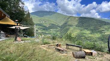 A peaceful outdoor setting with benches, logs, and a hammock, overlooking lush green mountains under a partly cloudy blue sky and surrounded by tall grass.