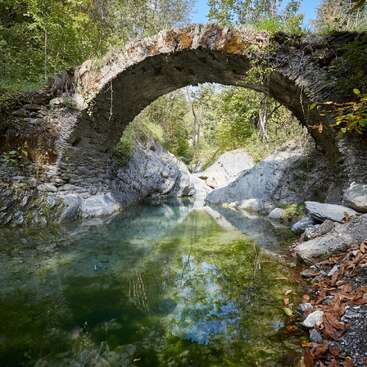 A stone arch bridge crosses over a serene, clear stream surrounded by rocks, green trees, and natural vegetation under a bright blue sunny sky.