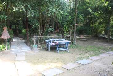 Um cenário de jardim tranquilo com um banco e uma mesa de madeira, cercado por árvores e vegetação exuberantes. Caminhos de pedra levam à tranquila área florestal próxima.